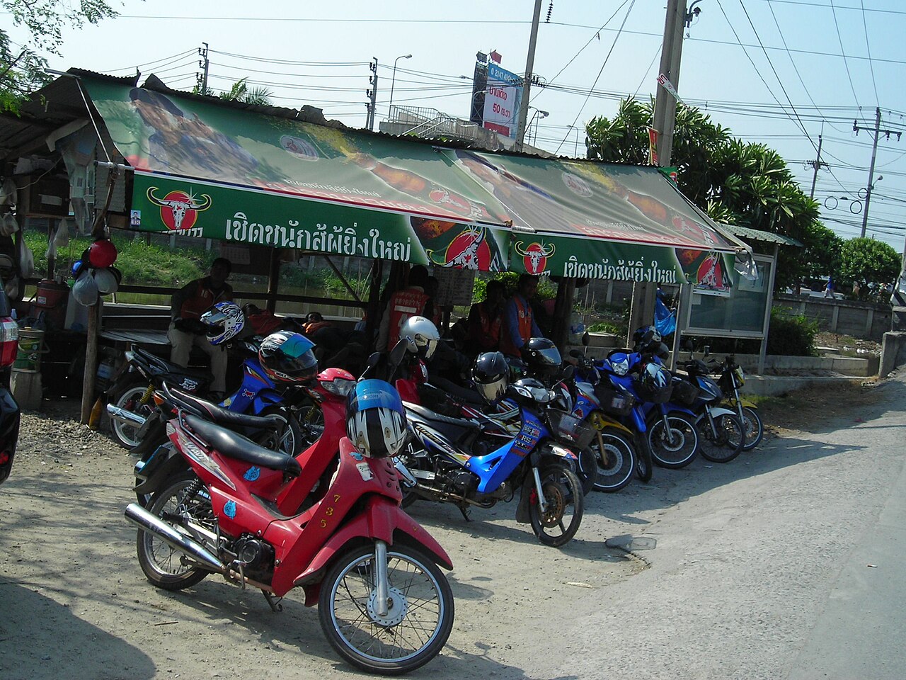 Motorbike taxi riders in numbered orange vests waiting with their bikes at a roadside stand near Bangkok, the 20 to 40 baht option that keeps Thailand travel costs down on short trips