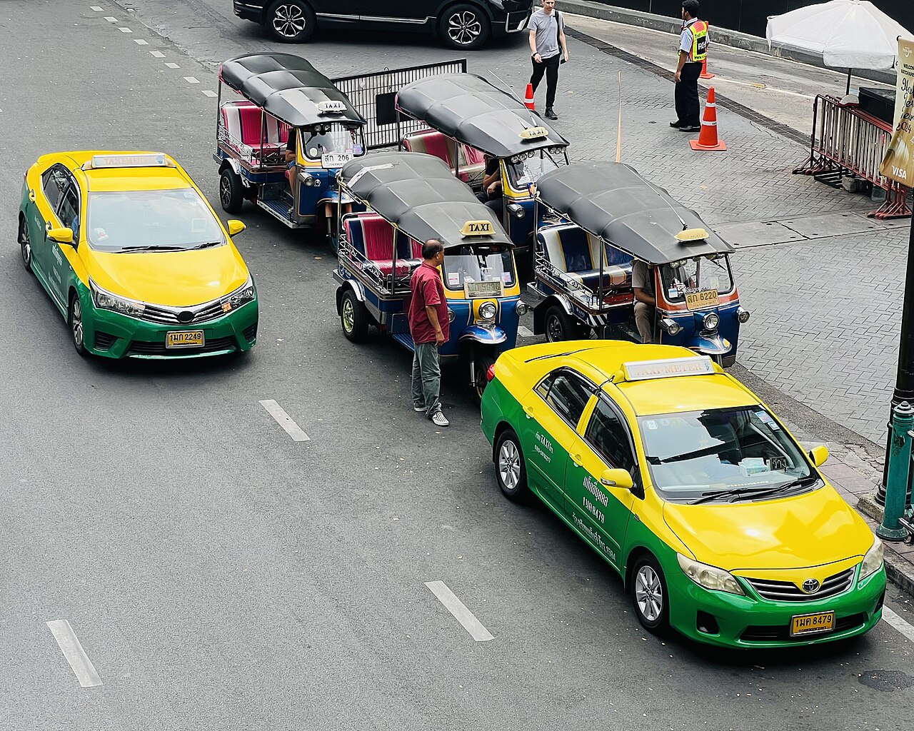Green and yellow metered taxis parked beside a line of tuk-tuks on Ratchadamri Road in central Bangkok, the two road options that decide Thailand travel costs for visitors