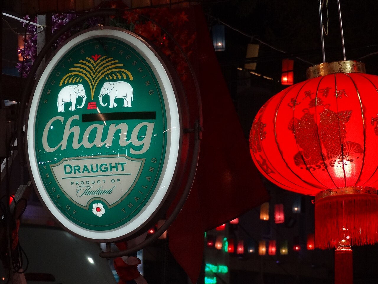 Illuminated Chang Draught bar sign hanging outside a Thai bar at night beside a red lantern, marking a venue where Thailand travel costs for a draught pint run 150 to 220 baht