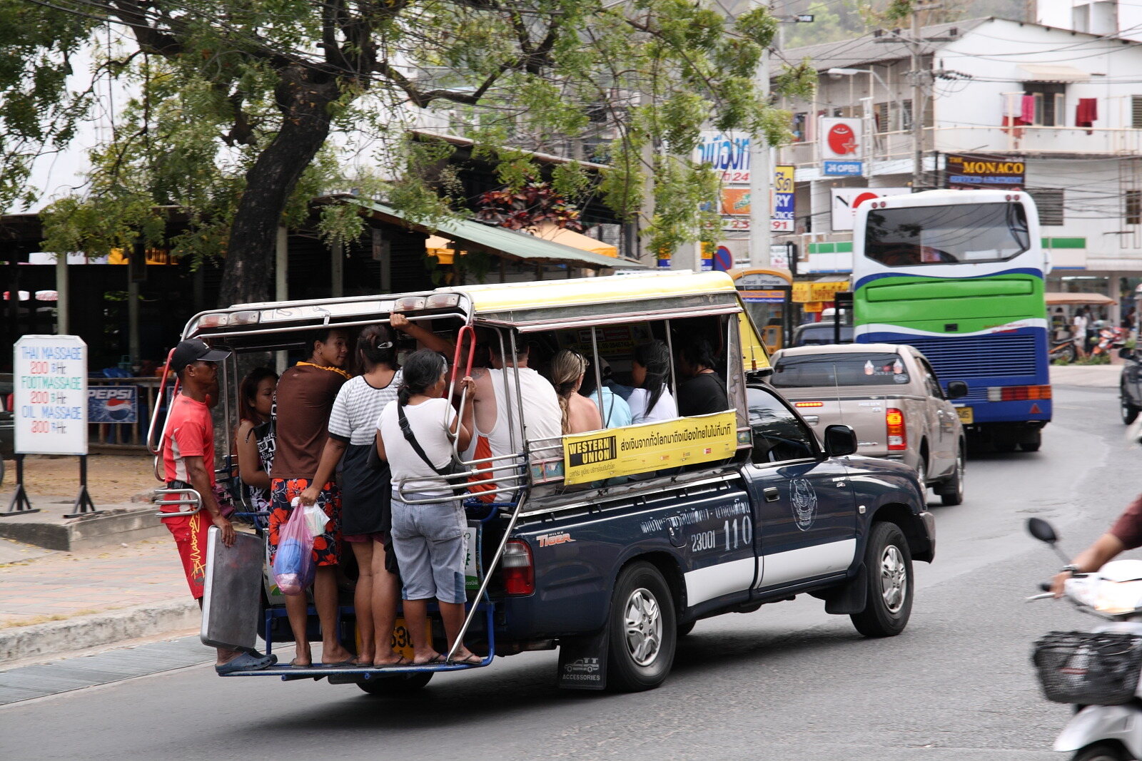 A crowded blue songthaew baht bus carrying standing passengers along Second Road in Pattaya, Thailand, where Thailand travel costs for a shared ride are 15 baht against 100 to 300 baht for a private charter