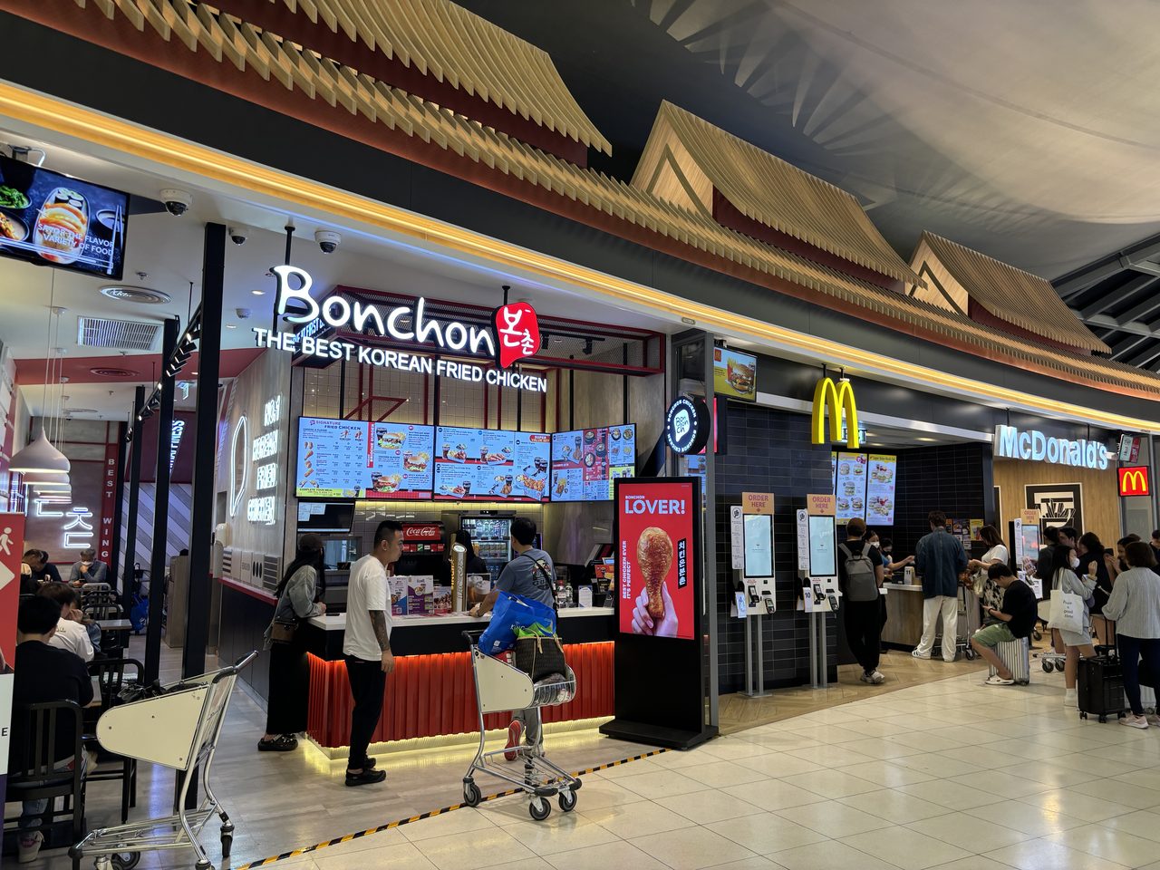 Bonchon and McDonald's outlets with lit menu boards and luggage trolleys inside the Suvarnabhumi Airport terminal in Bangkok, where Thailand travel costs for airport food run highest